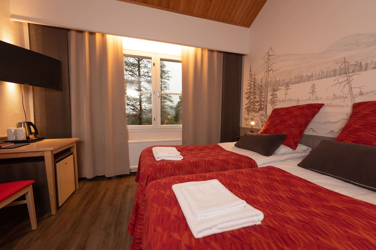 Habitación de hotel con dos camas, un escritorio, un televisor y ventana con vistas a árboles y montañas.