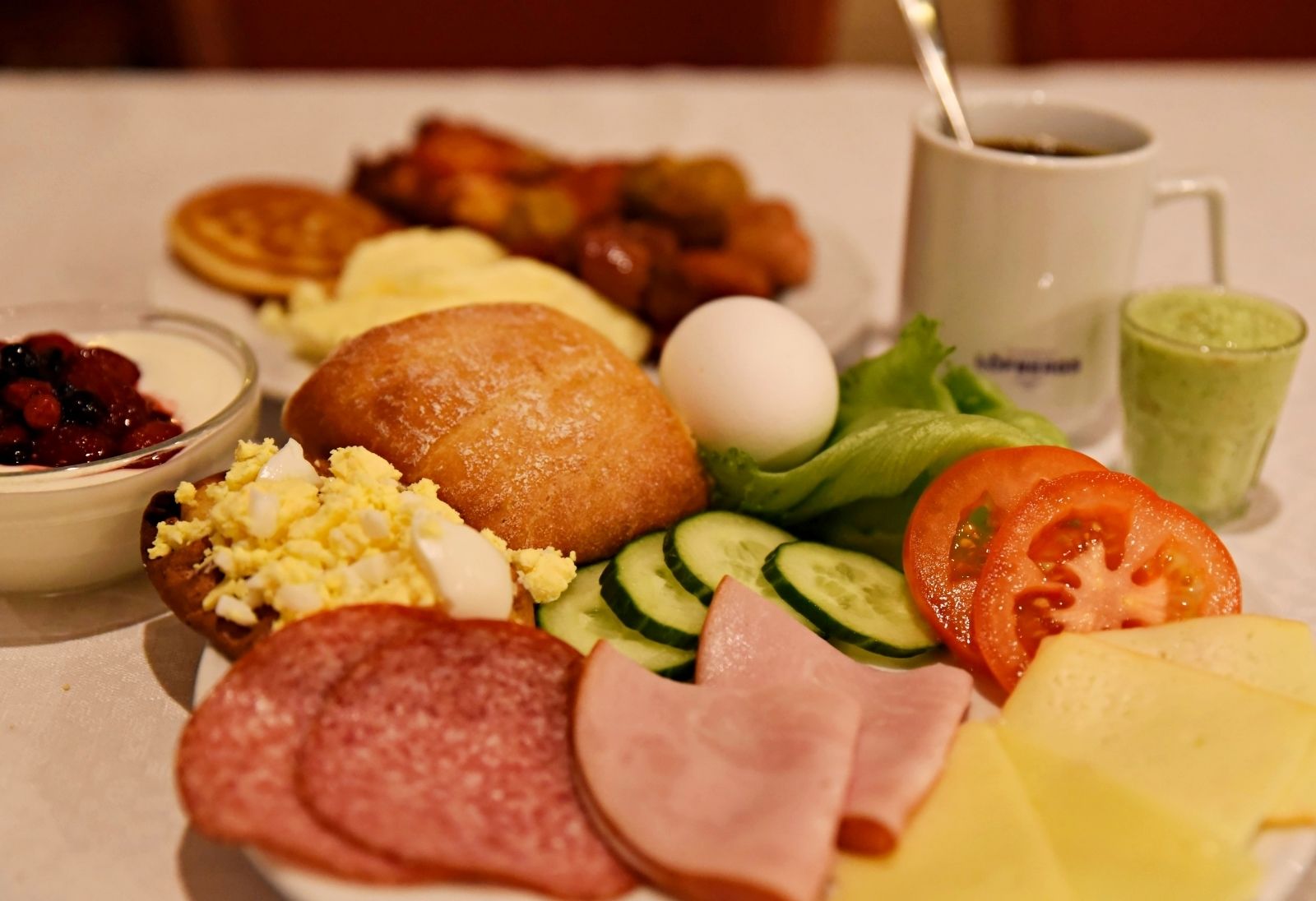 A breakfast plate with eggs, sausages, ham, cheese, tomatoes, cucumbers, and a cup of coffee.