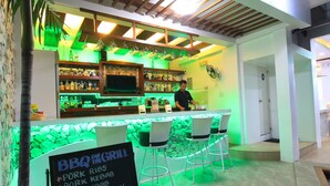 Un espace bar avec un tableau affichant le menu, proposant des plats de barbecue comme des travers de porc et des steaks, un barman et un comptoir de bar éclairé en vert.