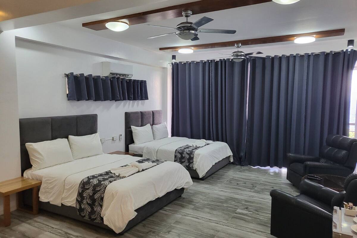 A hotel room with two beds, a ceiling fan, a desk, a chair, and a large window with curtains.