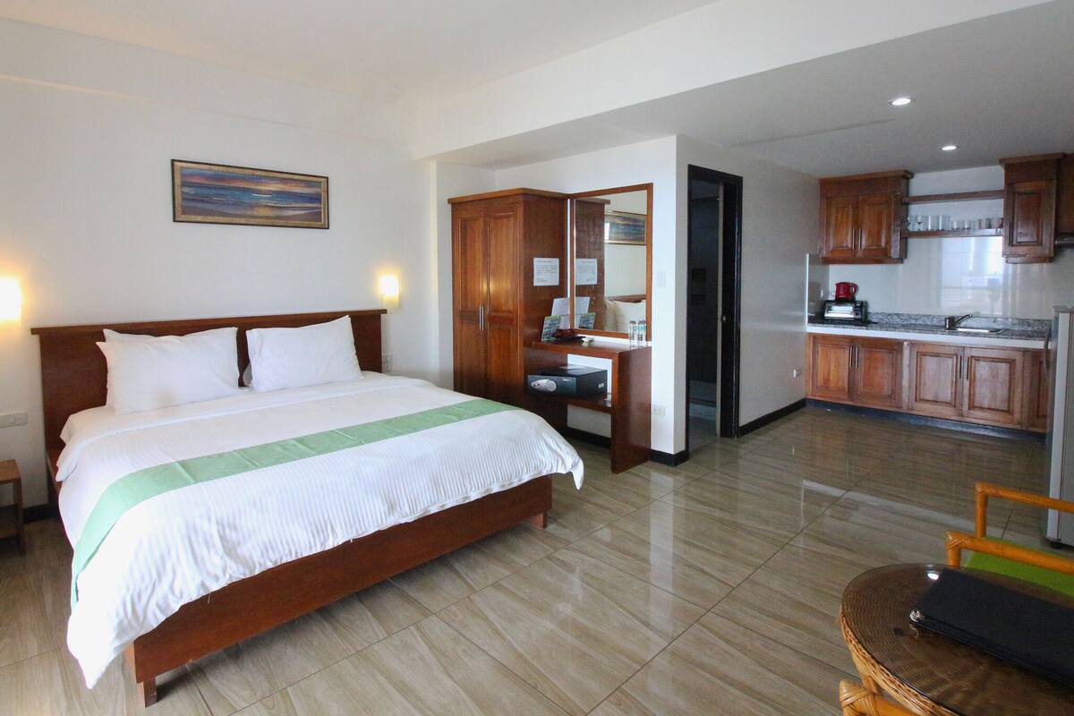 A hotel room with a large bed, a desk, a mirror, and a kitchenette.
