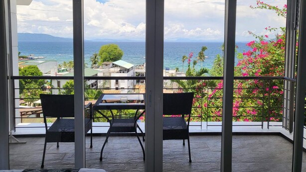 Un balcon avec une table et des chaises, une vue sur la mer et les bâtiments, et un arbuste fleuri.