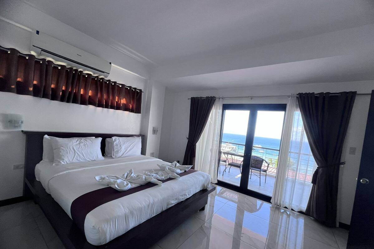 A hotel room with a large bed, a view of the ocean, and a balcony with a table and chairs.