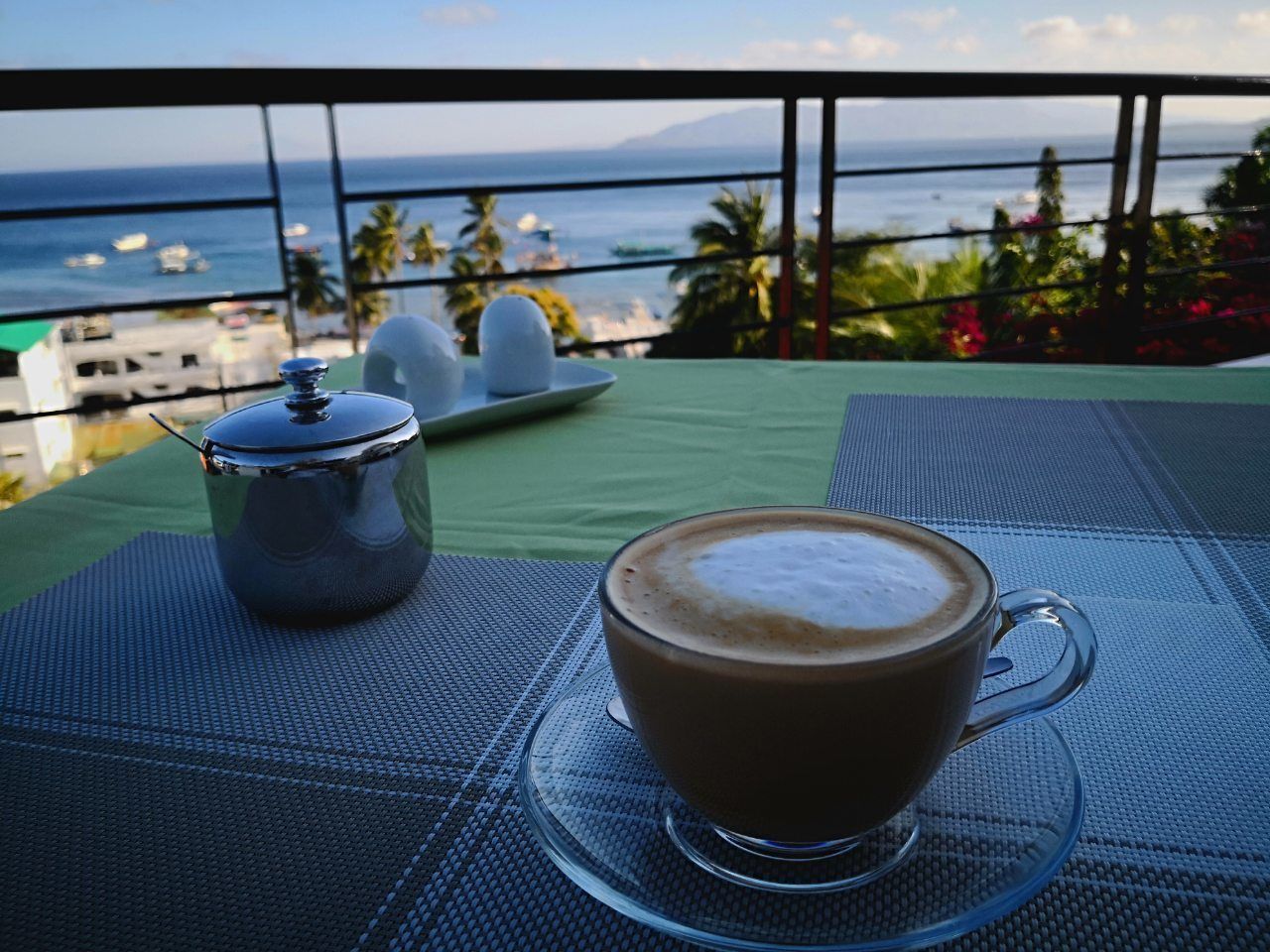 A cup of coffee on a table with a view of the ocean and palm trees.
