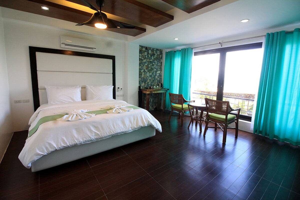 A hotel room with a large bed, a dining table with chairs, a view of the outdoors, and a ceiling fan.