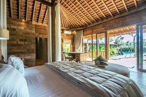 A bedroom with a large bed, a wooden ceiling, a desk, and a view of a pool.