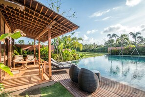A wooden deck with lounge chairs and a small table, overlooking a swimming pool with a view of palm trees and a garden.