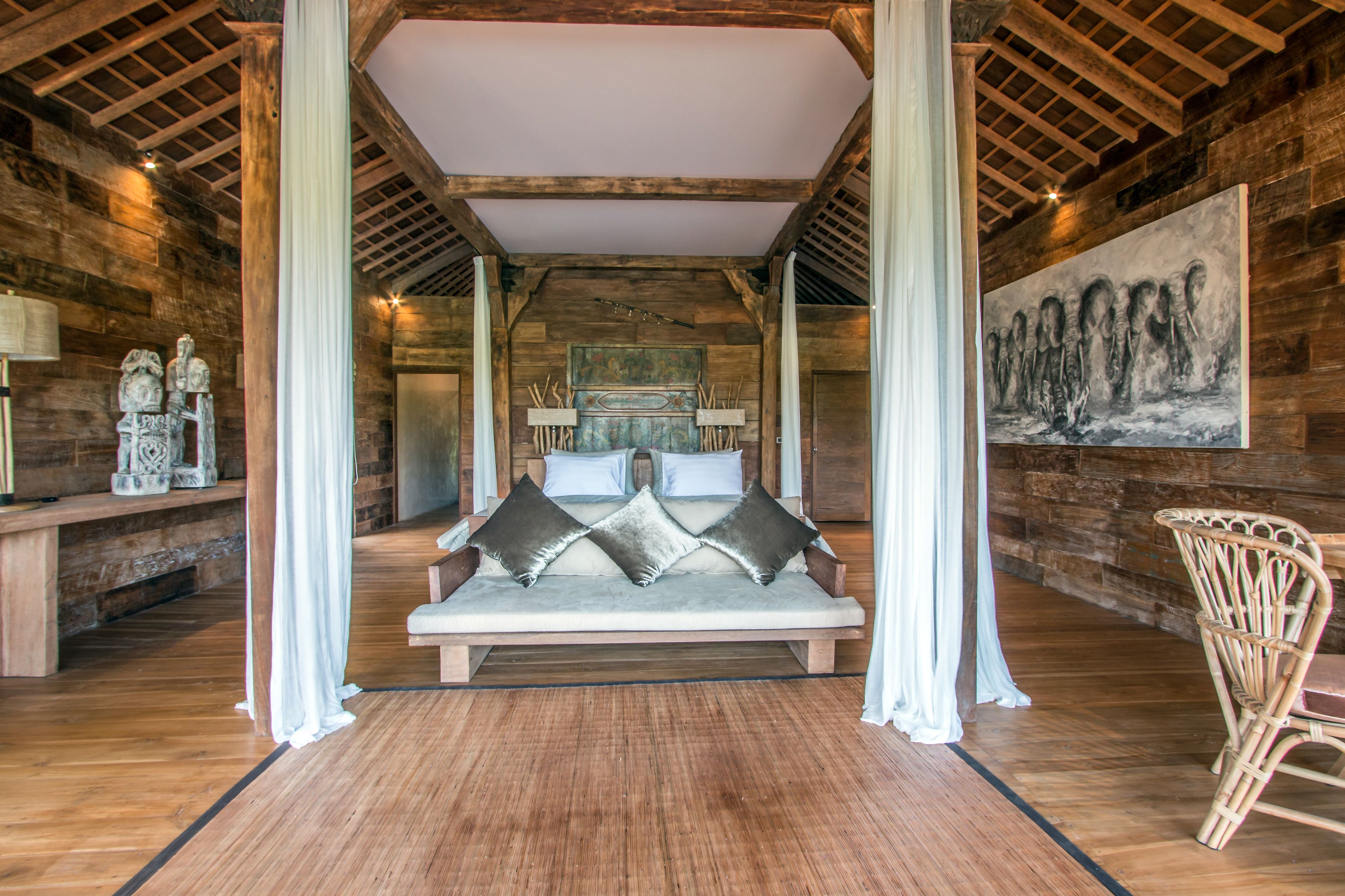 A cozy room with a wooden ceiling, a bed with pillows, a wicker chair, and a painting on the wall.