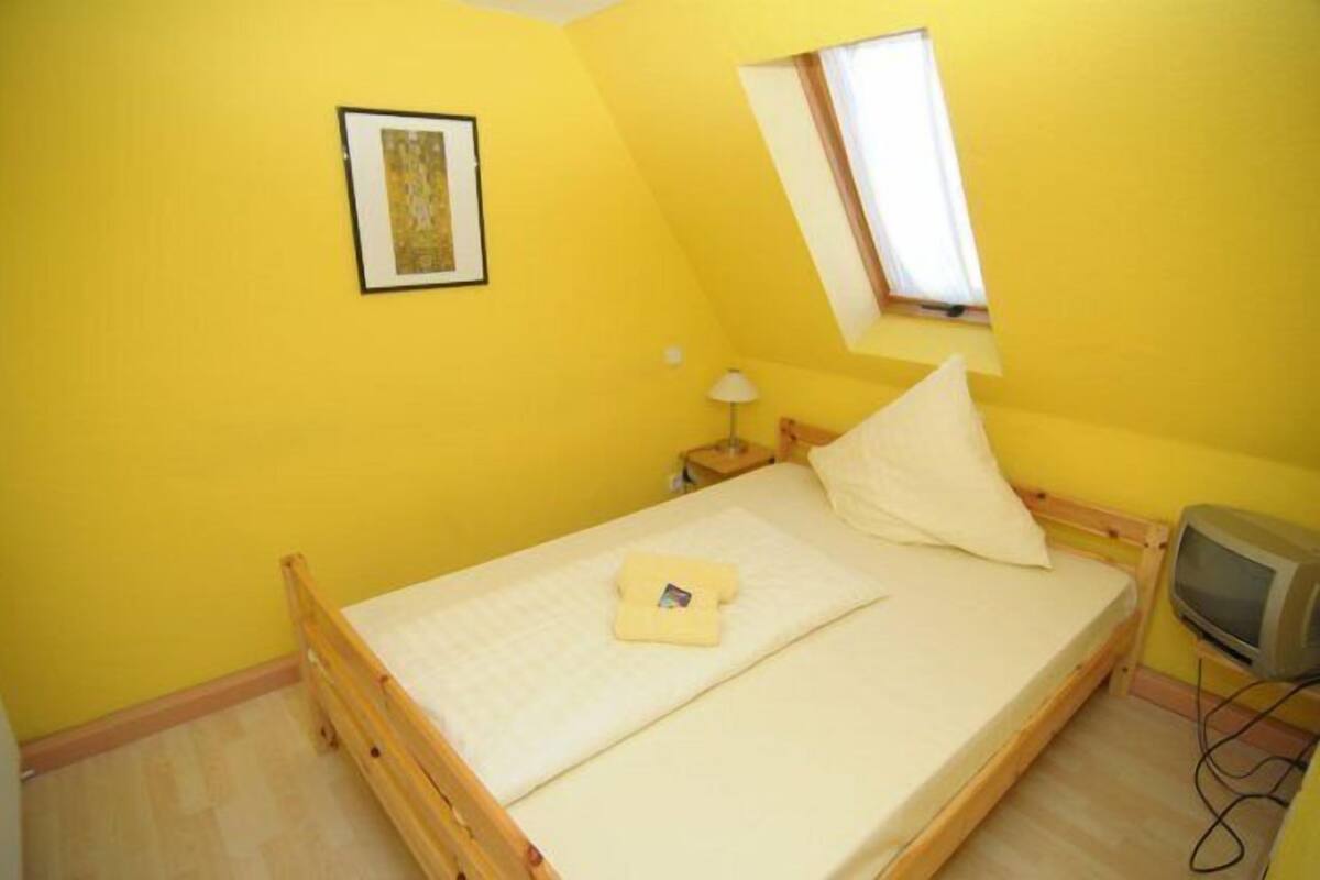 Una habitación pequeña y luminosa de color amarillo, con una cama individual, una lámpara de noche, un televisor y un cuadro en la pared.
