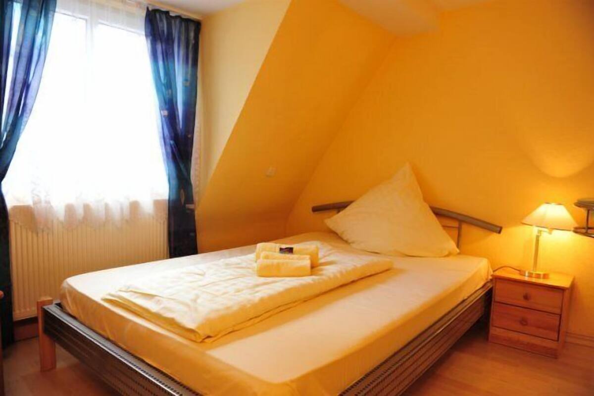 Una cama individual con edredón blanco, una mesita de noche con lámpara y una ventana con cortinas azules.