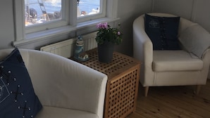 A room with a view of a dock and boats, a white sofa, a small wooden table with a plant, and a chair with a cushion. - Hostel Lotsen (Dalaro)
