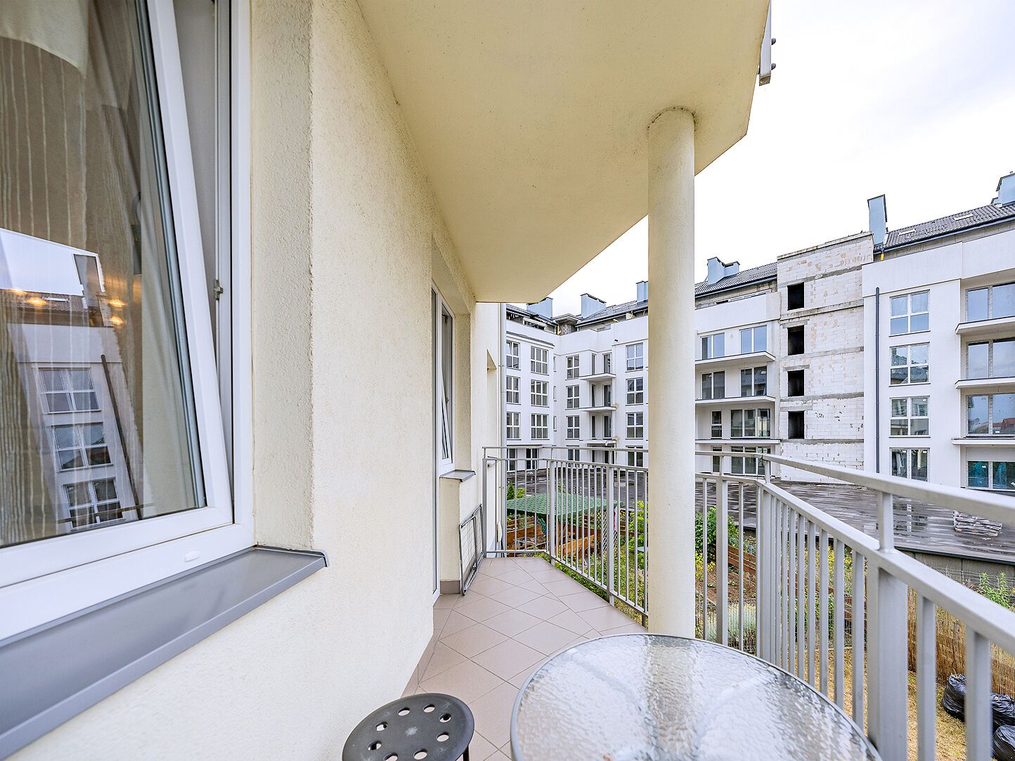 A balcony with a glass table and a chair, overlooking a residential area with multiple buildings.