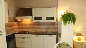 Cocina compacta con alacenas blancas, estufa a gas y un lavamanos. Hay una planta colgante y una mesita de noche con lámpara.