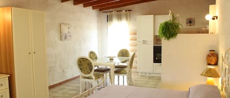 A bedroom with a bed, a wardrobe, a dining table with chairs, a kitchen area, and a hanging plant.