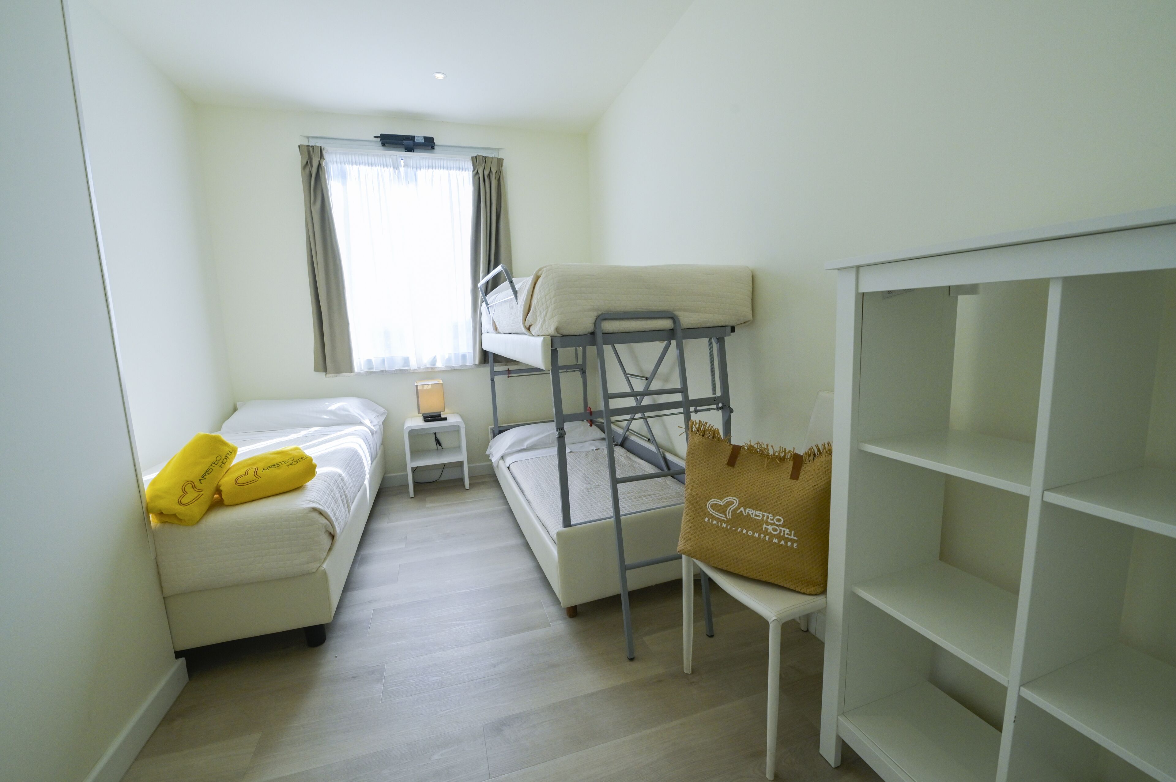 Habitación de hotel compacta con una litera, una mesita de noche pequeña, una lámpara y una estantería.