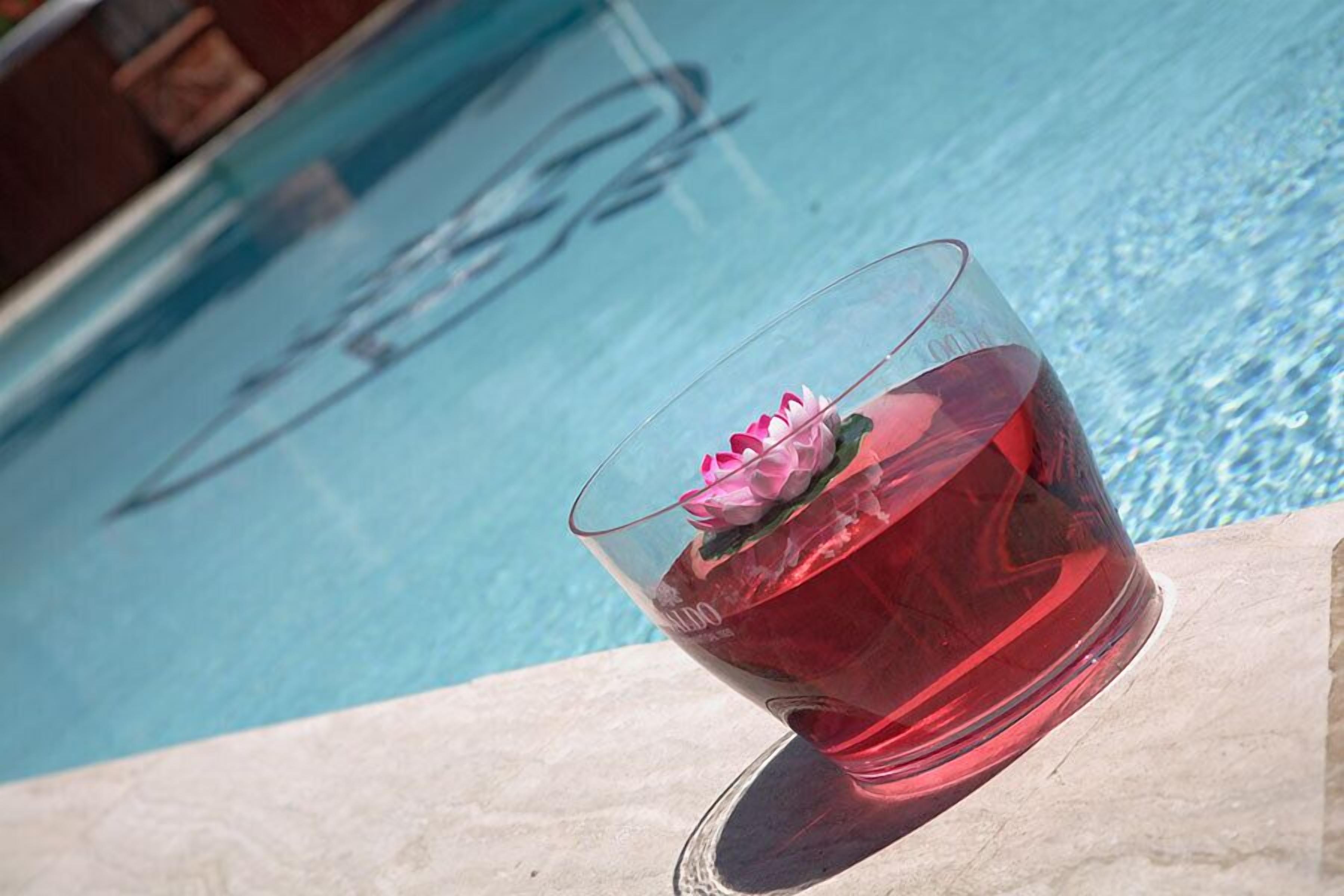 Un verre contenant une fleur rose et un liquide rouge, placé près d’une piscine.