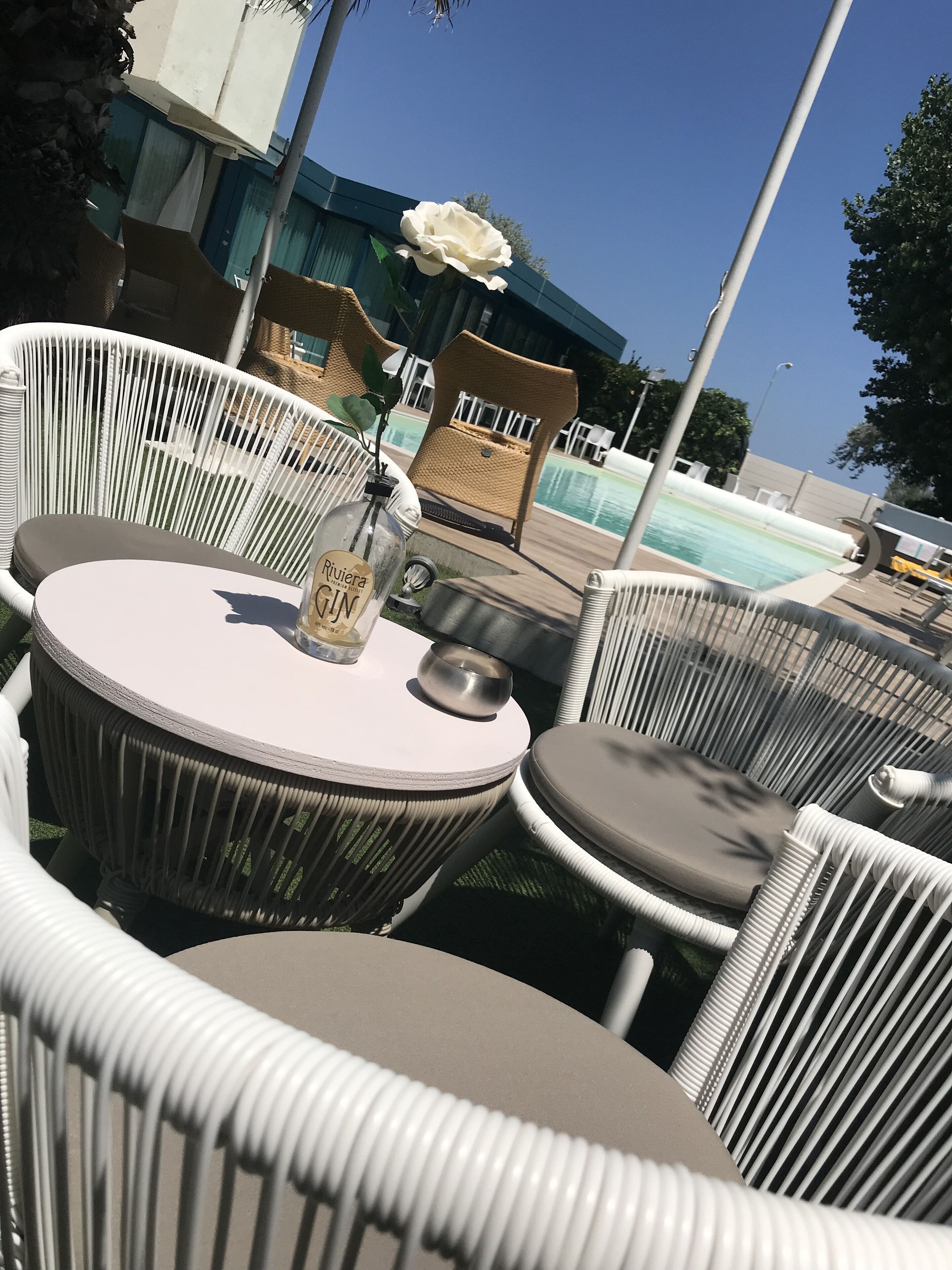 Zona junto a la piscina con sillas de mimbre y una mesa redonda, una botella de ginebra y una flor blanca.