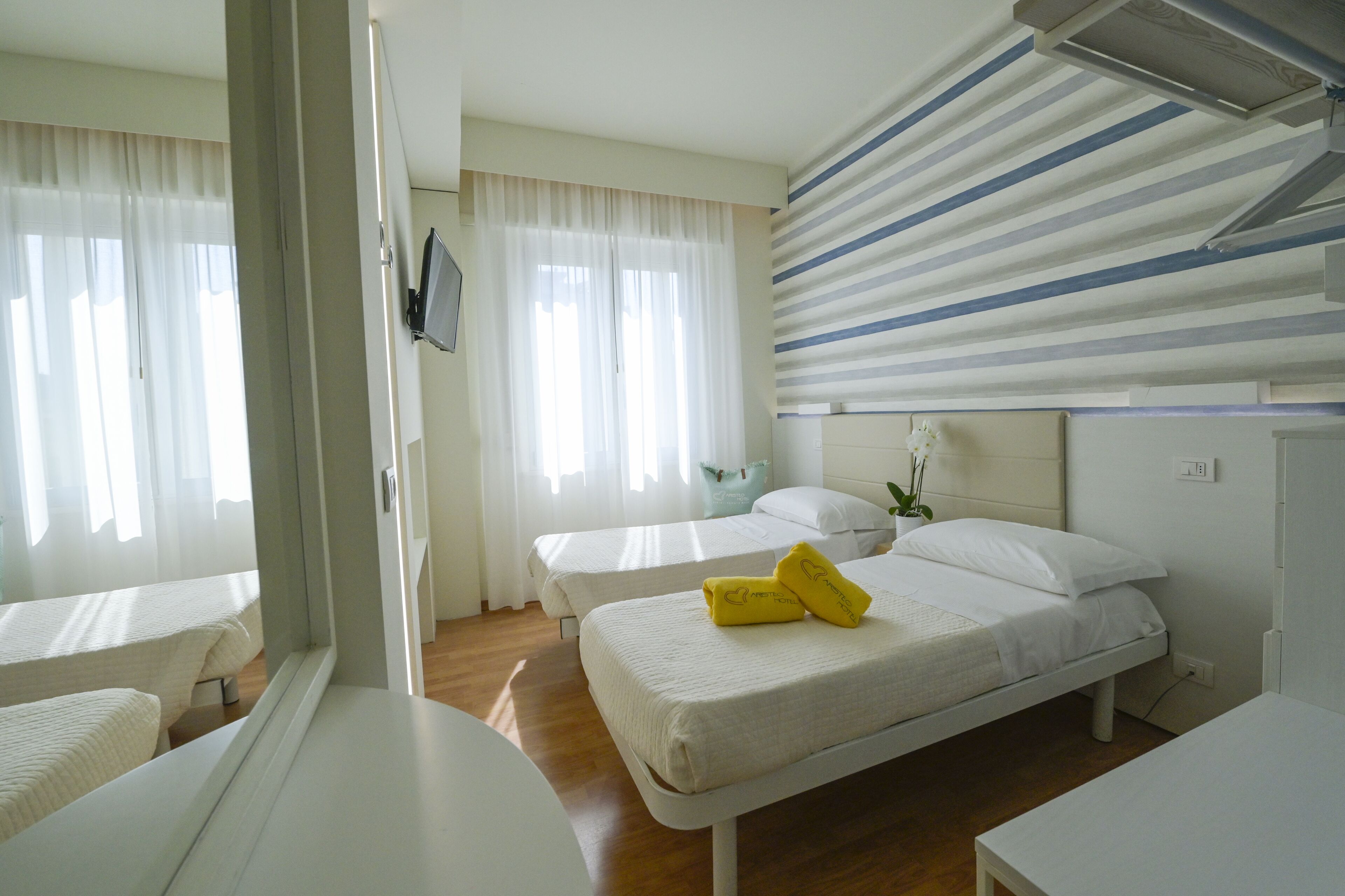 A hotel room with two beds, a large window with curtains, a flat-screen TV mounted on the wall, and a striped ceiling.