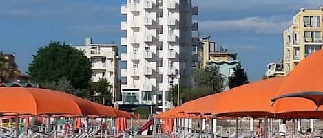 A beach with rows of sun loungers under orange umbrellas, a modern white building in the background, and other residential buildings.