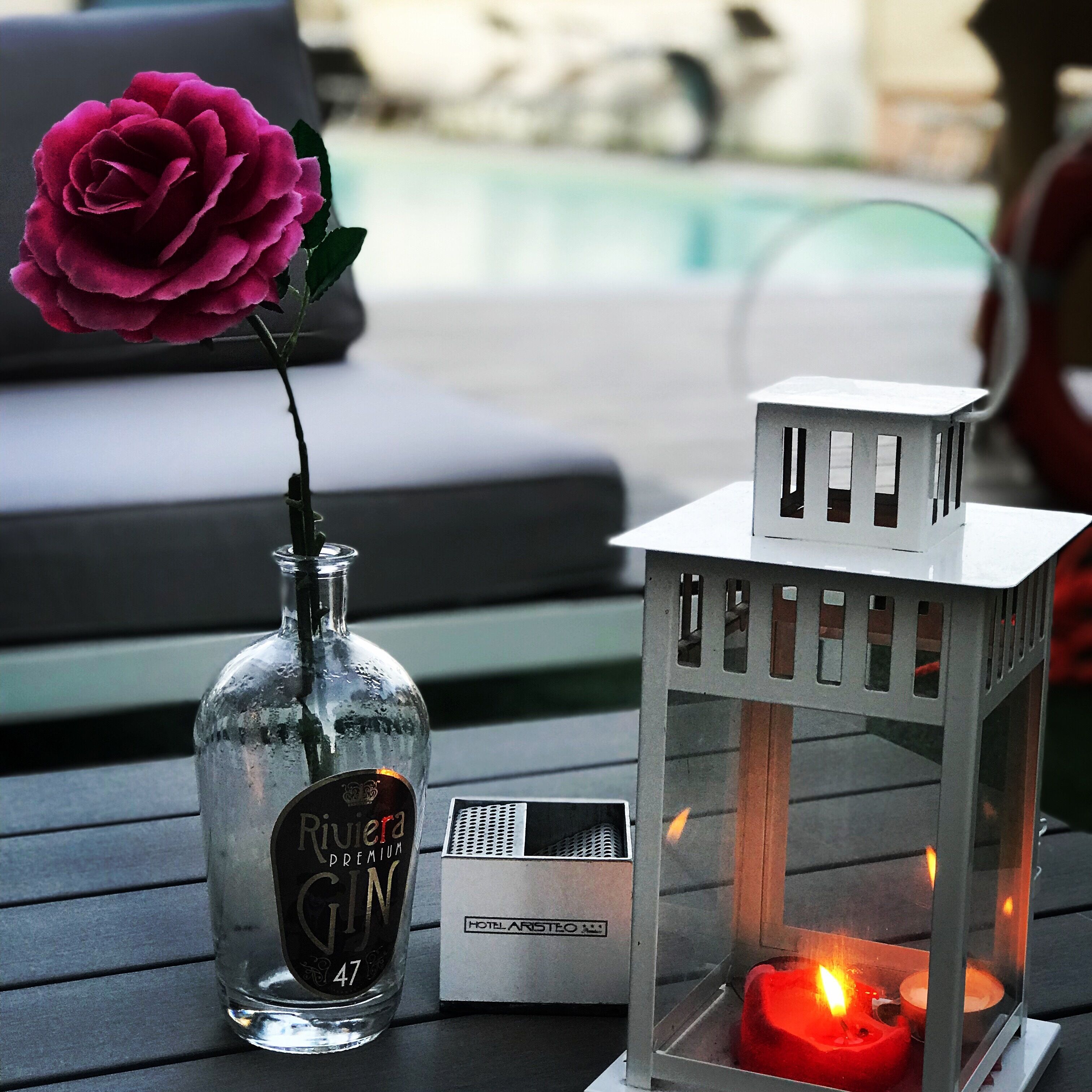 Une bouteille de gin Rivera Premium en verre, une rose rouge, une lanterne blanche et une boîte d’allumettes sur une table.