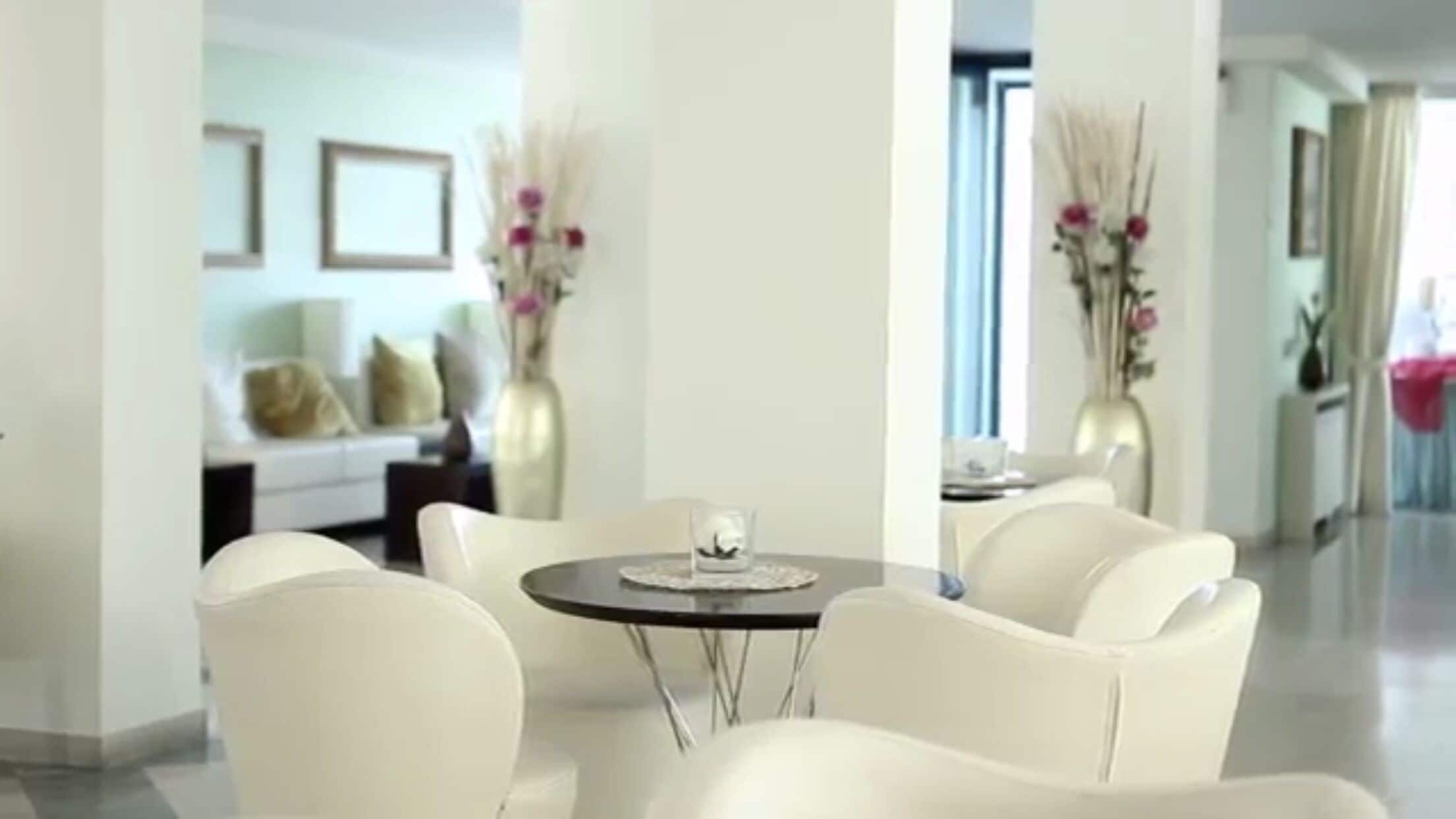 A modern hotel lobby with a round table and four white chairs, a sofa in the background, and two vases with flowers.