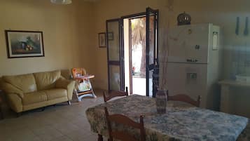 Uma sala de jantar com mesa e cadeiras, um sofá, uma geladeira e uma porta que dá para o exterior.