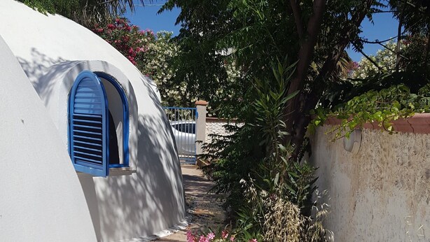 Uma passagem estreita ladeada por paredes brancas e janelas com venezianas azuis, que leva a um edifício com uma porta visível.