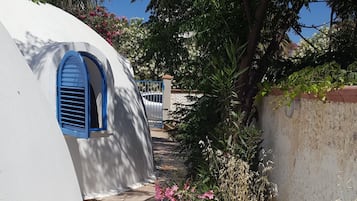 Uma passagem estreita ladeada por paredes brancas e janelas com venezianas azuis, que leva a um edifício com uma porta visível.