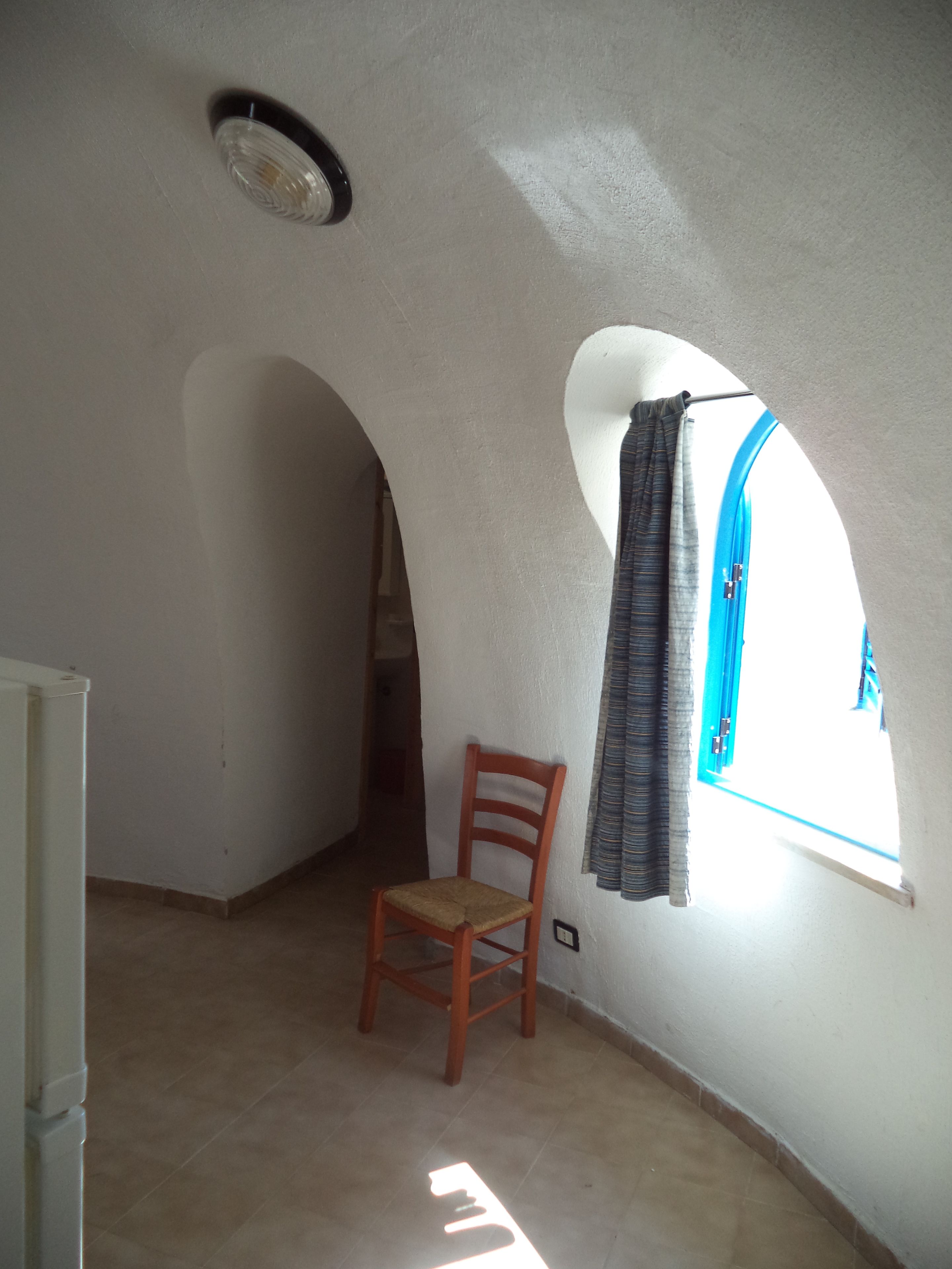 Quarto com teto alto em arco, uma cadeira de madeira, janela com cortinas e luminária visível.