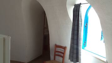 Quarto com teto alto em arco, uma cadeira de madeira, janela com cortinas e luminária visível.