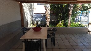 A patio with a table and chairs, a red bowl, and a view of a garden with trees and a blue gate. - Bungalow Tonnarella Beach (Mazara del Vallo)