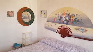 Un dormitorio con cama, mesita de noche, espejo, ventilador de pared y un cuadro con motivos florales.
