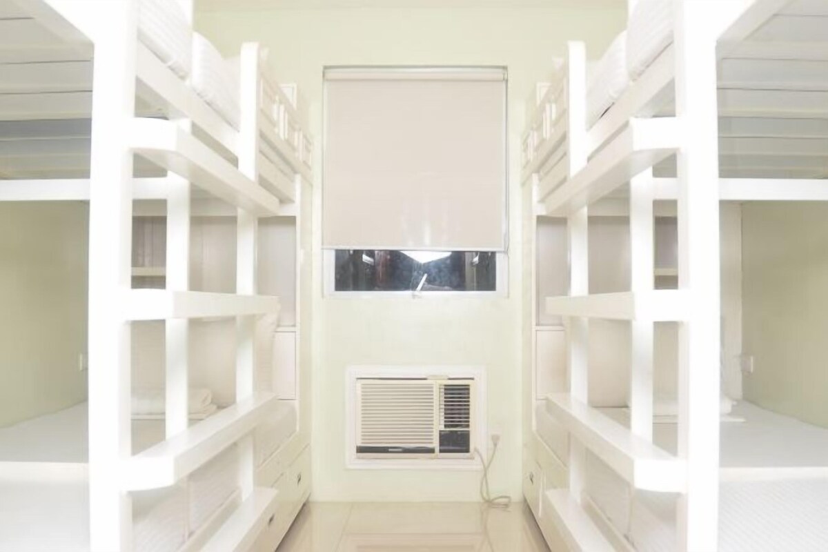 Habitación con literas dispuestas en un pasillo, una ventana con persianas y un aire acondicionado de pared.