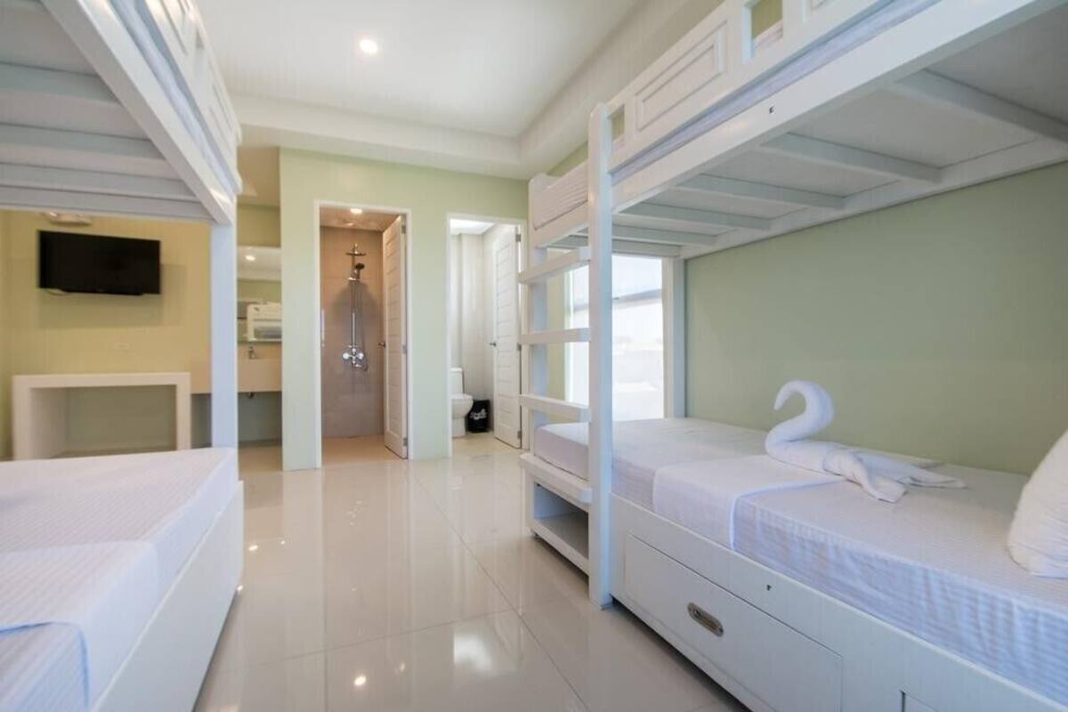 A hotel room with bunk beds, a TV, a bathroom with a shower, and a hallway leading to another room.