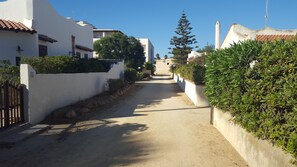 A narrow, unpaved street flanked by white buildings with red-tiled roofs, surrounded by greenery and a clear blue sky.