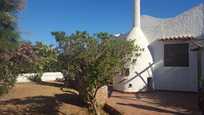 A white building with a unique conical roof, surrounded by a garden with flowering plants and a brick pathway.