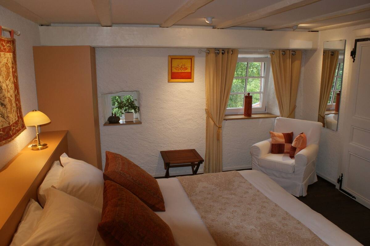 A bedroom with a bed, a chair, a nightstand, a lamp, a window with curtains, a small table, and a wall-mounted picture.