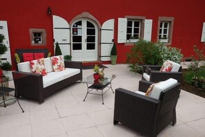 Un bâtiment rouge avec une porte et des volets blancs, meublé de canapés en osier et d’une table en verre.