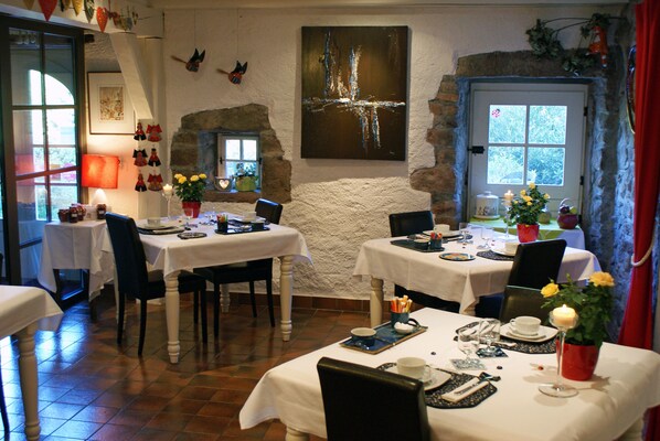 Un espace repas avec des tables préparées pour deux personnes, agrémenté de nappes blanches, de chaises et d’un mur en pierre rustique.