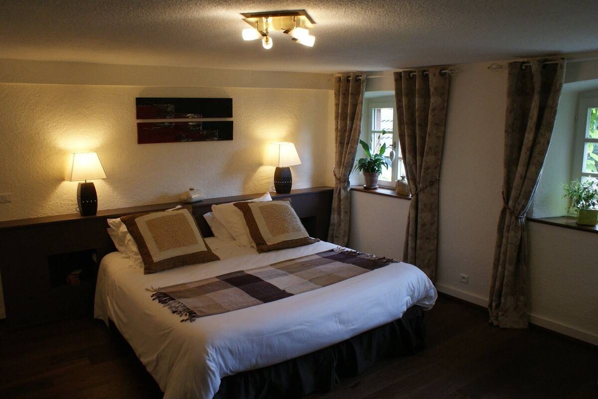A neatly made bed with a quilt, two bedside lamps, a window with curtains, and a wall-mounted artwork.