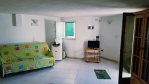 A small, sparsely furnished room with a green patterned sofa, a small table with a TV, and a window with green shutters.