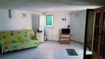 Una habitación pequeña y poco amoblada, con un sofá de color verde estampado, una mesa pequeña con un televisor y una ventana con contraventanas verdes.