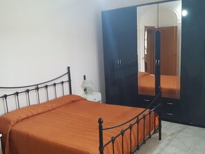 A bed with an orange bedspread, a white bedside table with a lamp, and a dark blue wardrobe with a mirror.