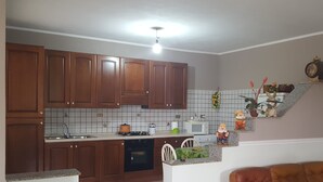 A kitchen with wooden cabinets, a microwave, a stove, and a sink. There is a chair and a small table with decorative items. - Villa Valeria (Mazara del Vallo)