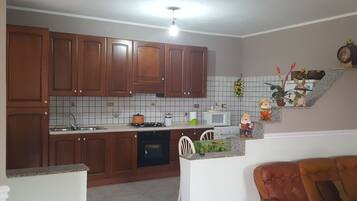 Una cocina con armarios de madera, microondas, estufa y lavabo. Hay una silla y una mesa pequeña con objetos decorativos.
