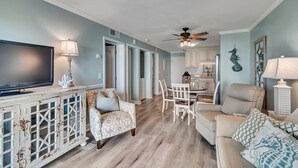Un salon avec un téléviseur, des fauteuils à motifs, un canapé, une table à manger avec des chaises, et une cuisine en arrière-plan.