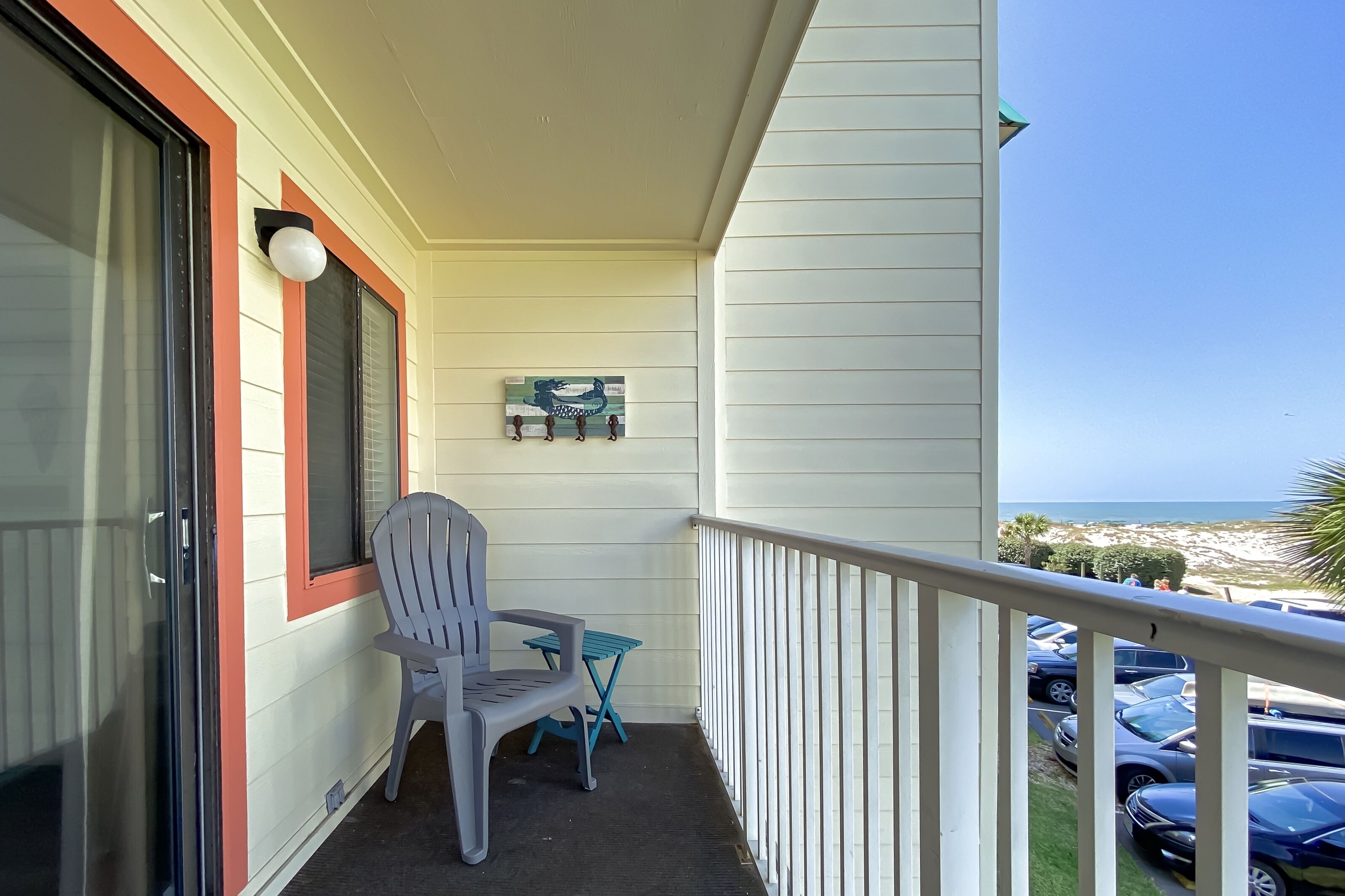 A balcony with a white chair and a small table, overlooking a parking area and beach.