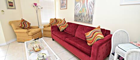 Una sala de estar con un sofá rojo, un sillón amarillo, una mesita de mimbre blanco y una mesa de centro de vidrio.