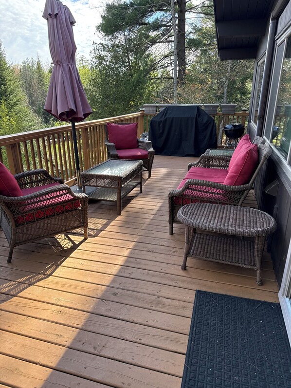Eine Holzterrasse mit Korbmöbeln, einem überdachten Grill und Blick auf Bäume.