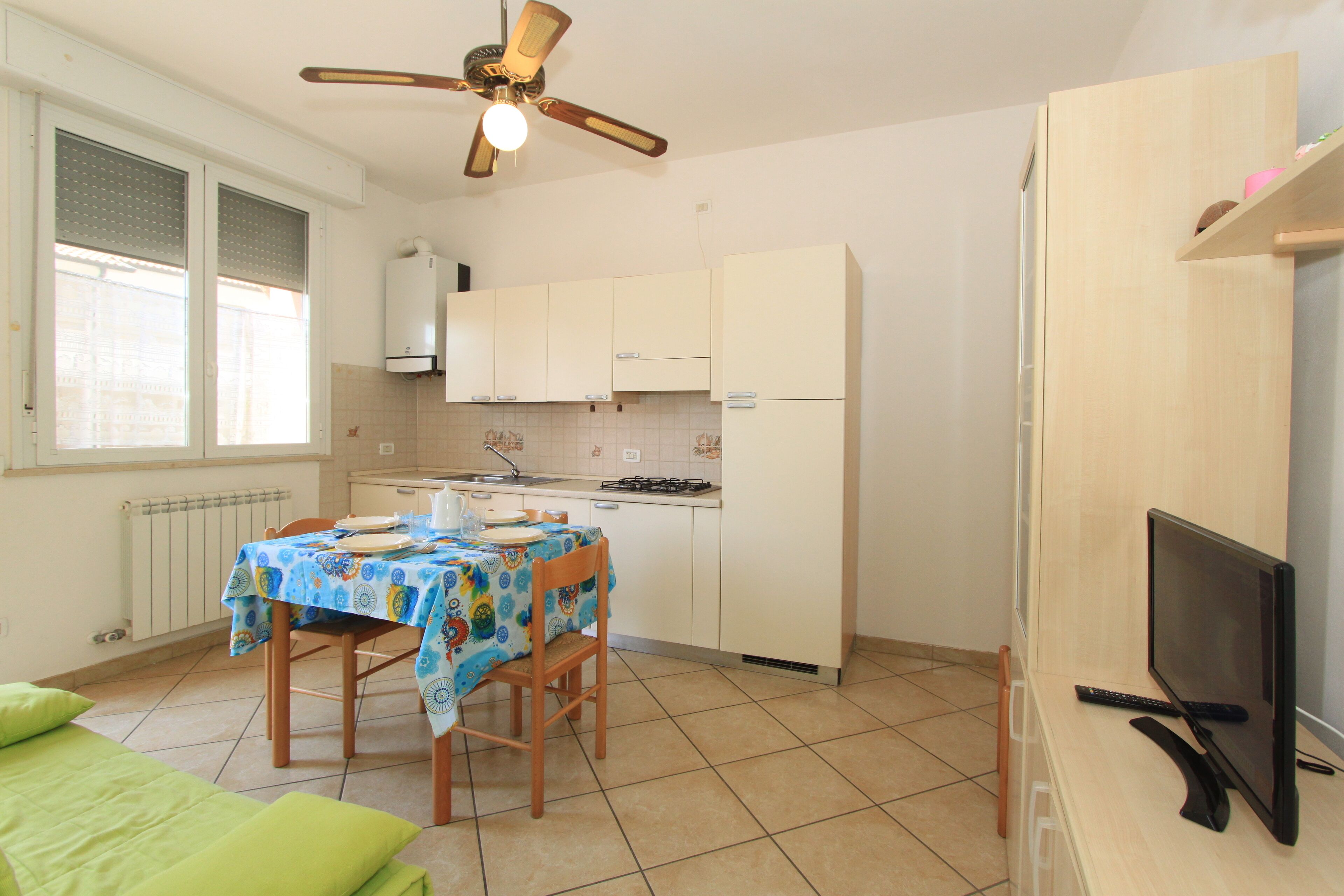 Una cocina compacta con una mesa de comedor, sillas, un ventilador de techo y un televisor sobre un mueble de madera.